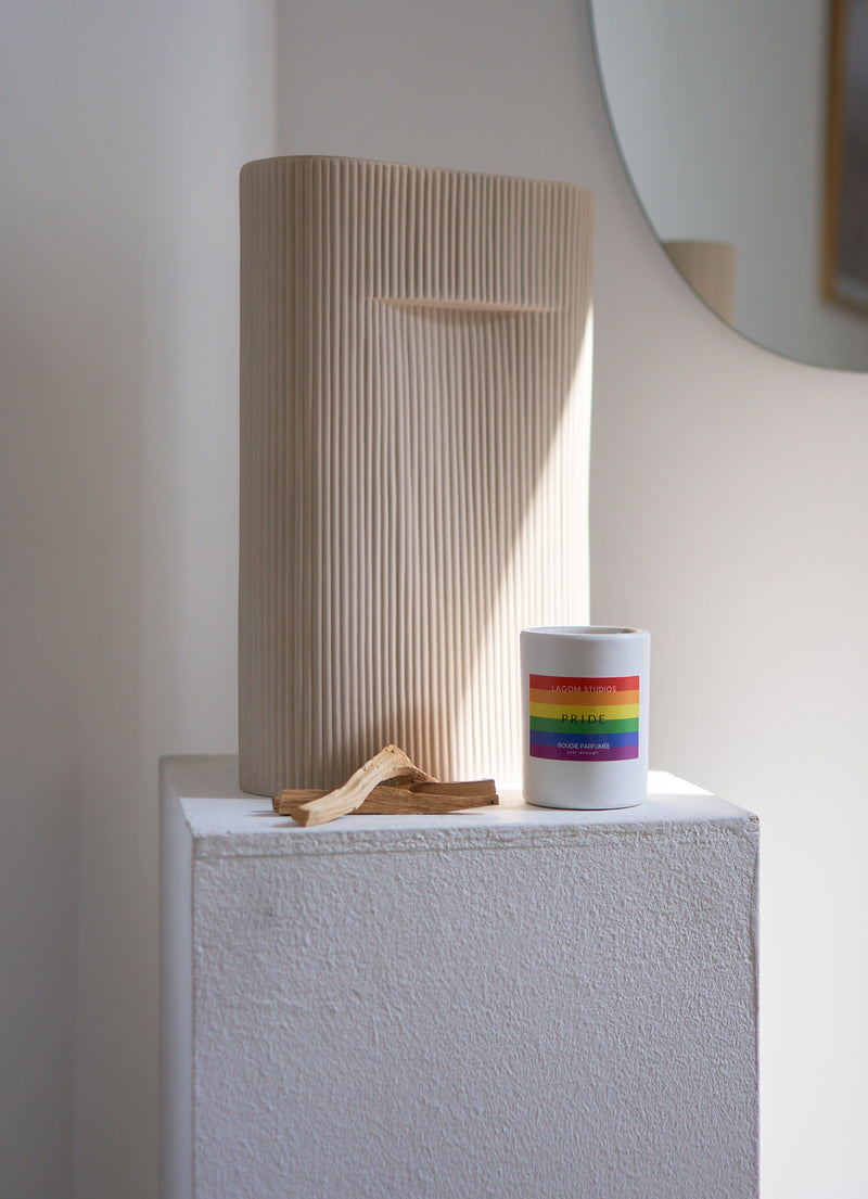 Weiße Duftkerze mit PRIDE Flagge auf Galeriesockel vor geriffelter Vase_Regenbogen Flagge